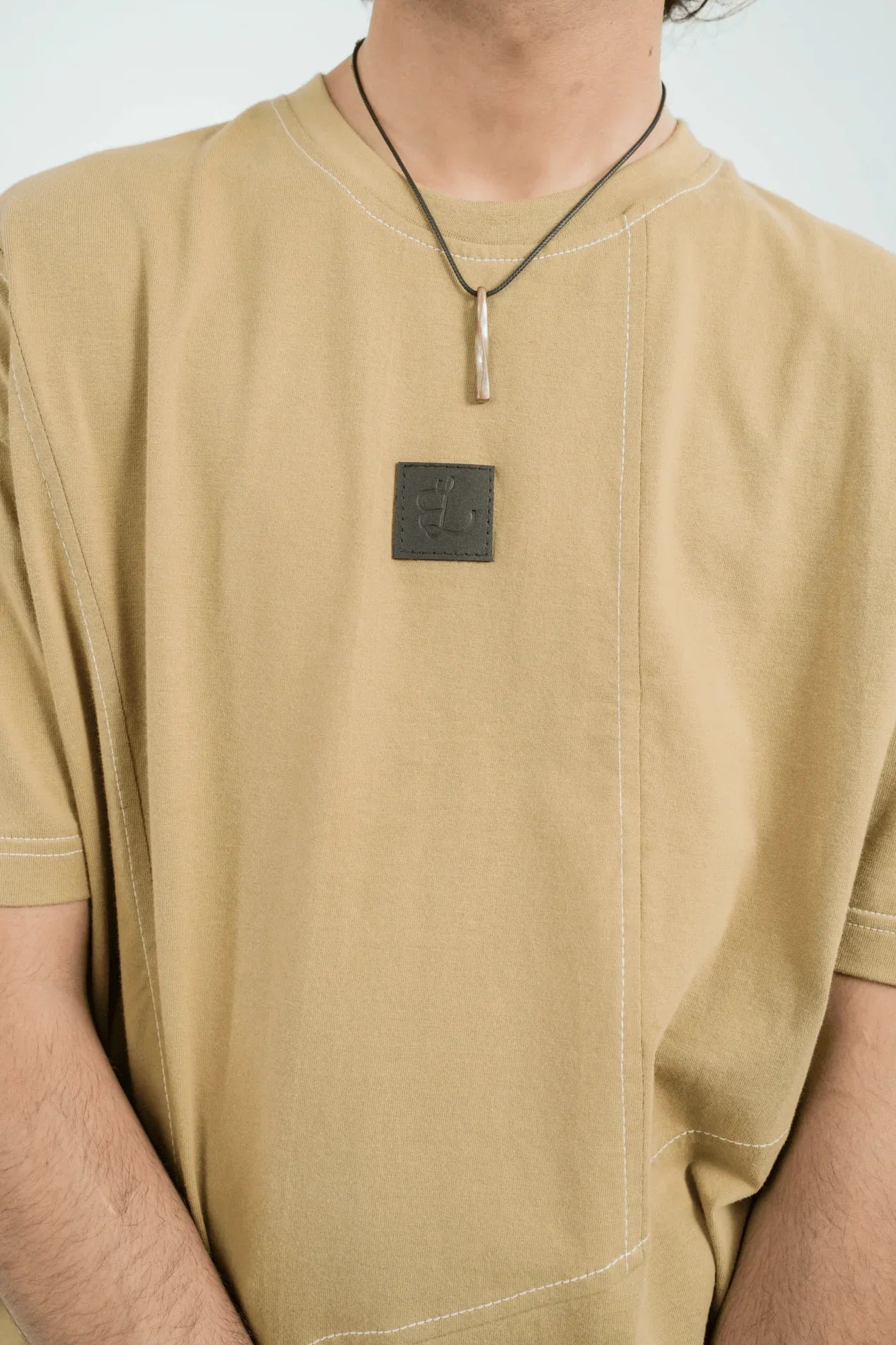 Close-up of person wearing beige t-shirt with black stitched logo patch and silver pendant necklace