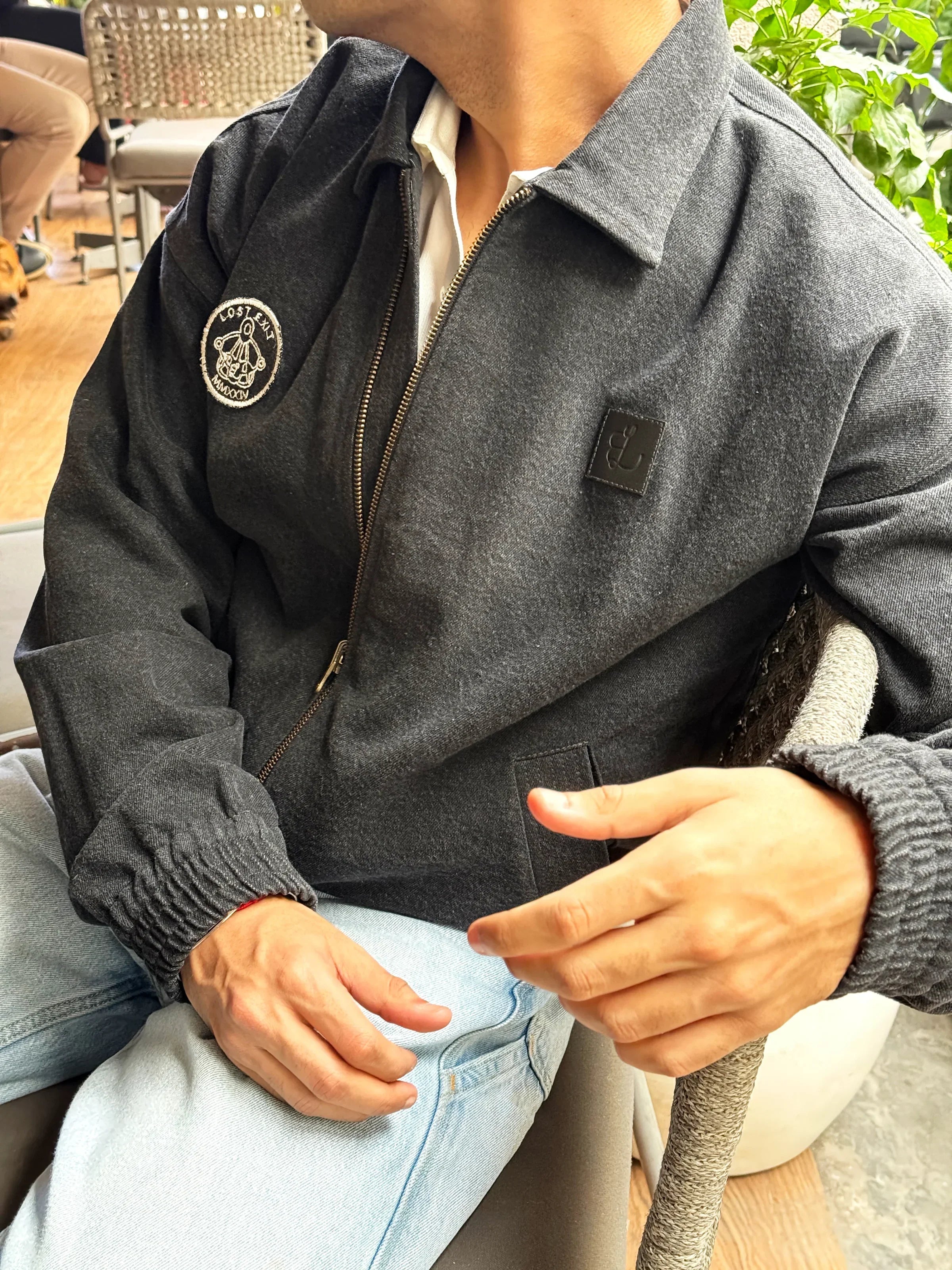 Man wearing charcoal gray zip-up jacket with patches, light blue jeans, sitting on woven chair indoors
