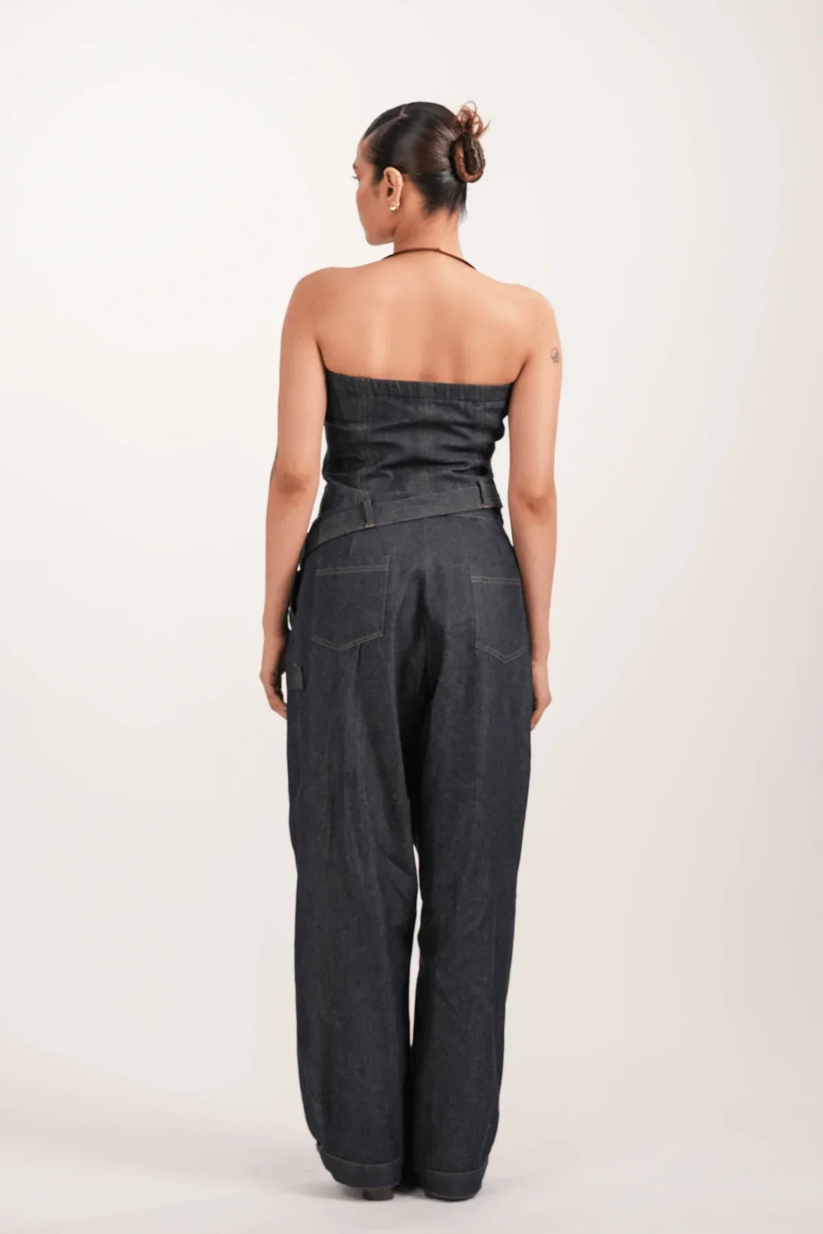 Rear view of woman wearing dark denim strapless jumpsuit with wide-leg pants against white background