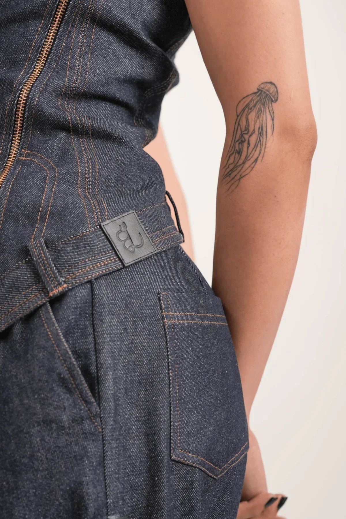 Close-up of a person wearing dark blue denim overalls with orange stitching and a jellyfish tattoo on their arm