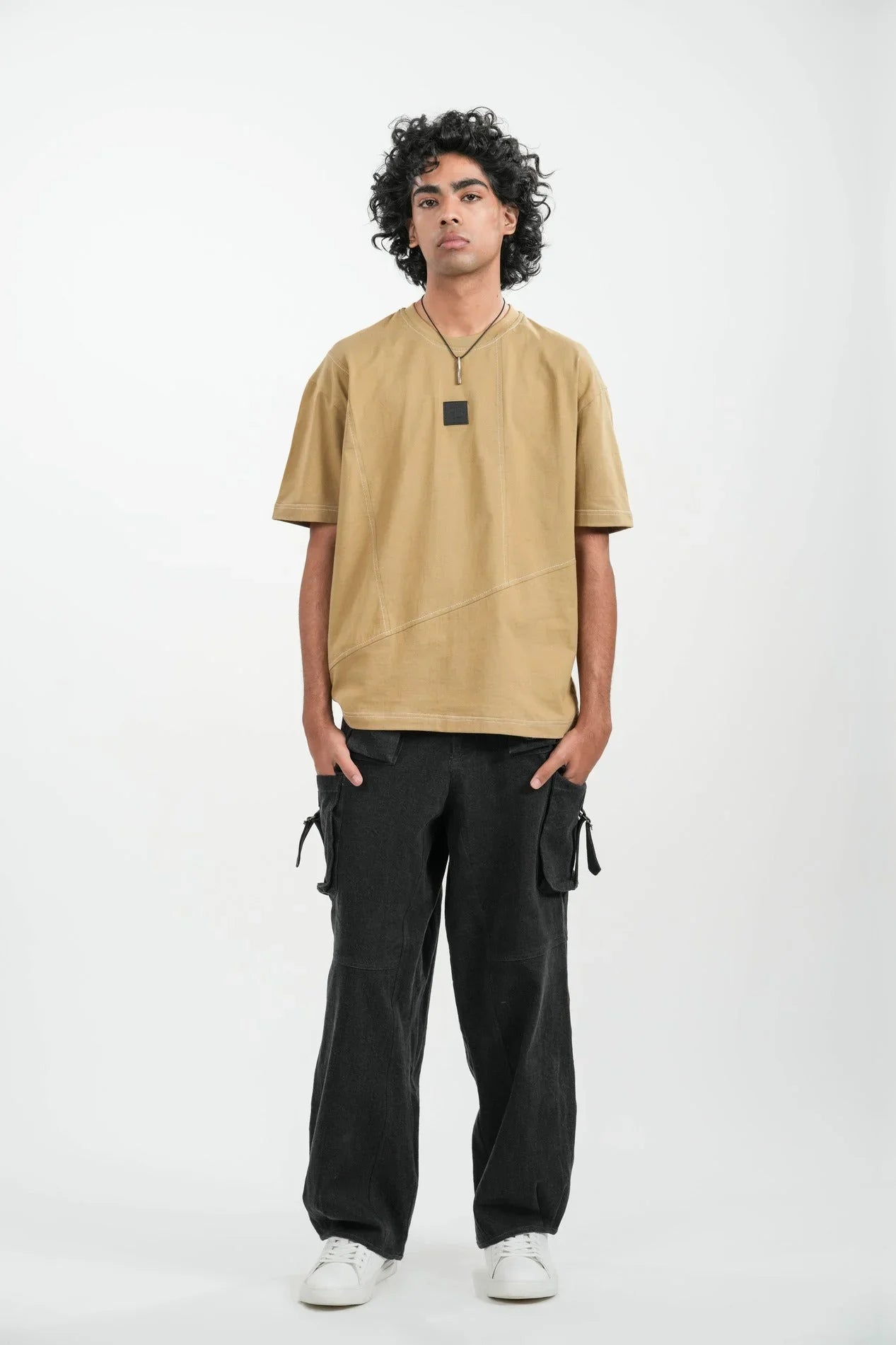 man wearing tan oversized t-shirt and black cargo pants with white sneakers standing against white background