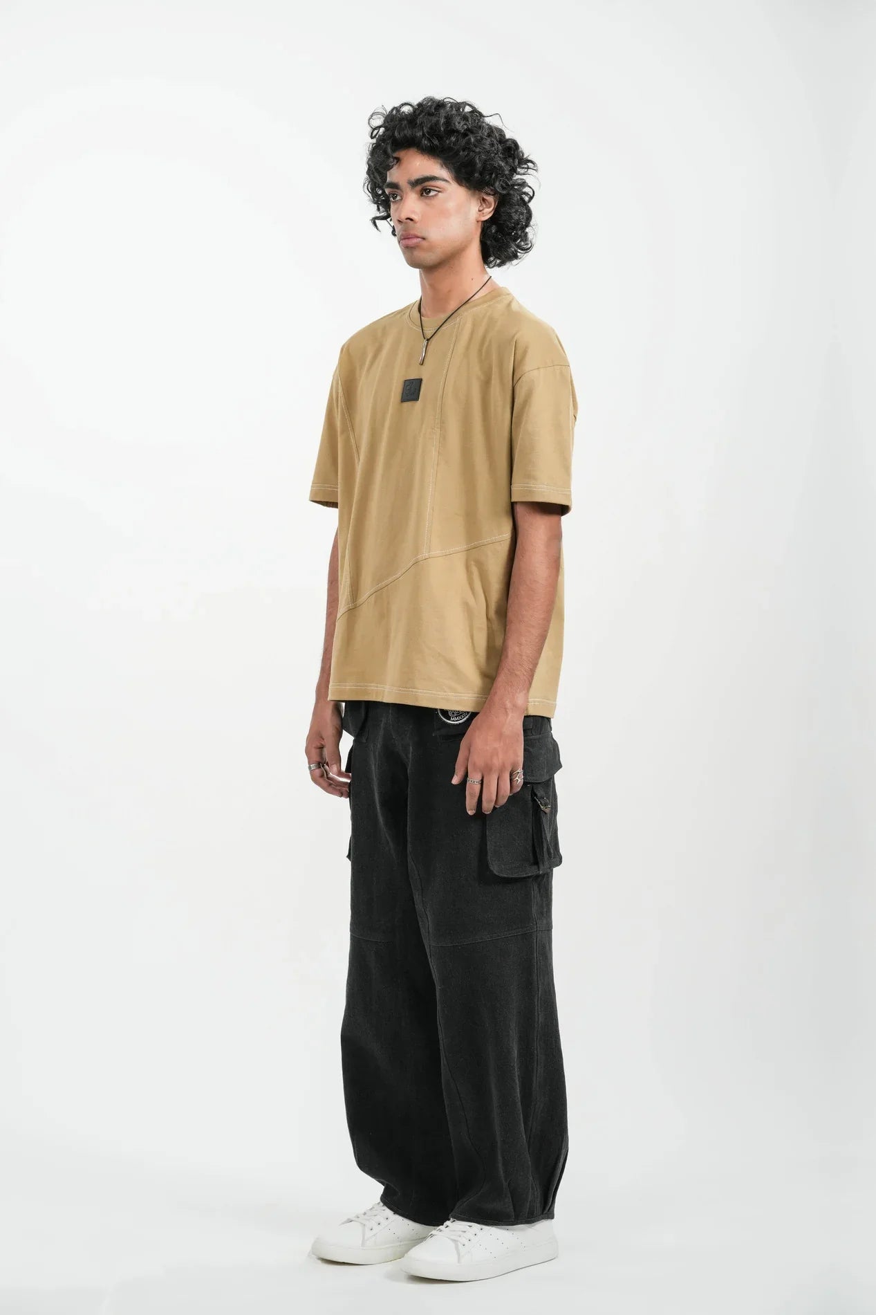 Young man wearing tan oversized t-shirt, black cargo pants, and white sneakers on white background