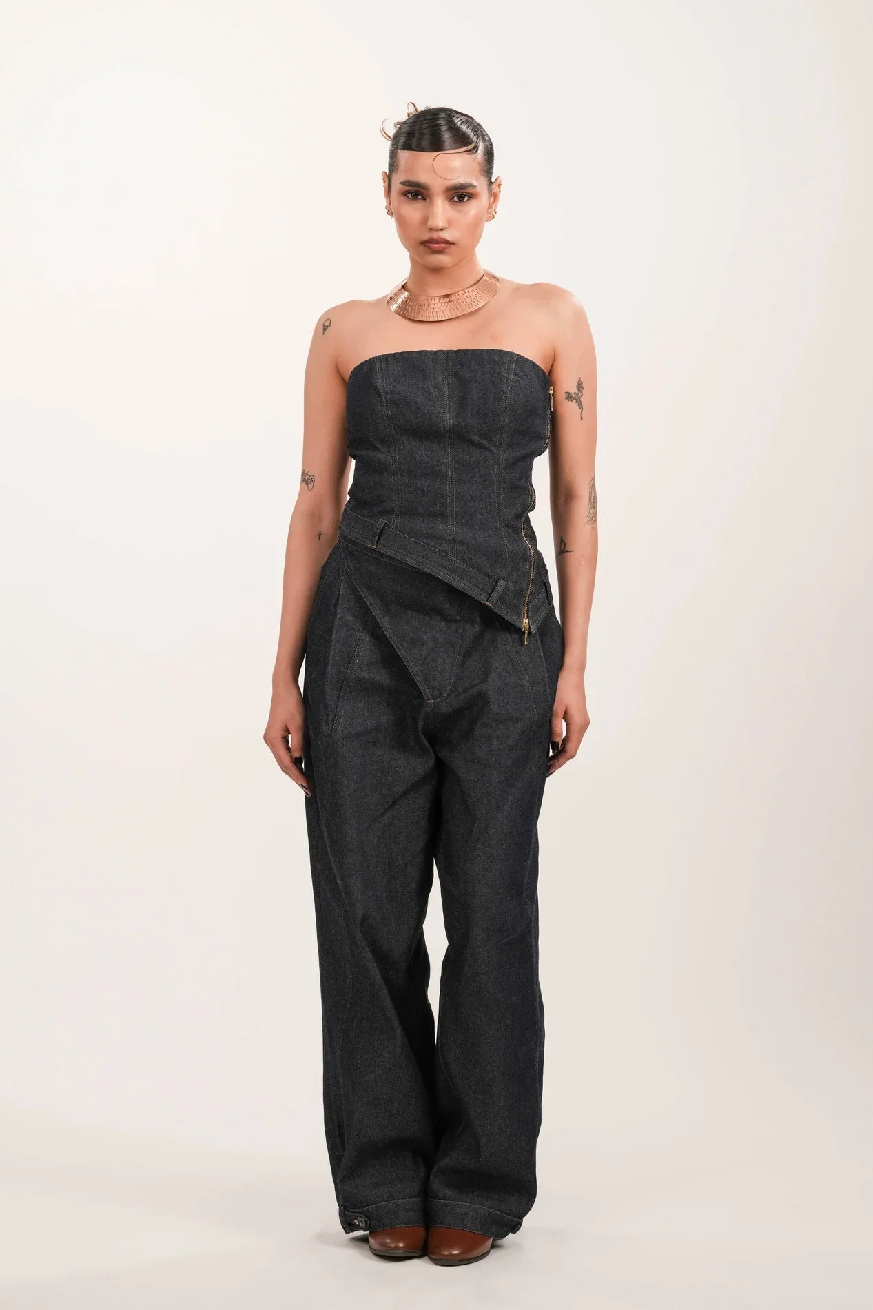 Model wearing strapless dark denim jumpsuit with wide legs, brown shoes, and copper choker against plain background