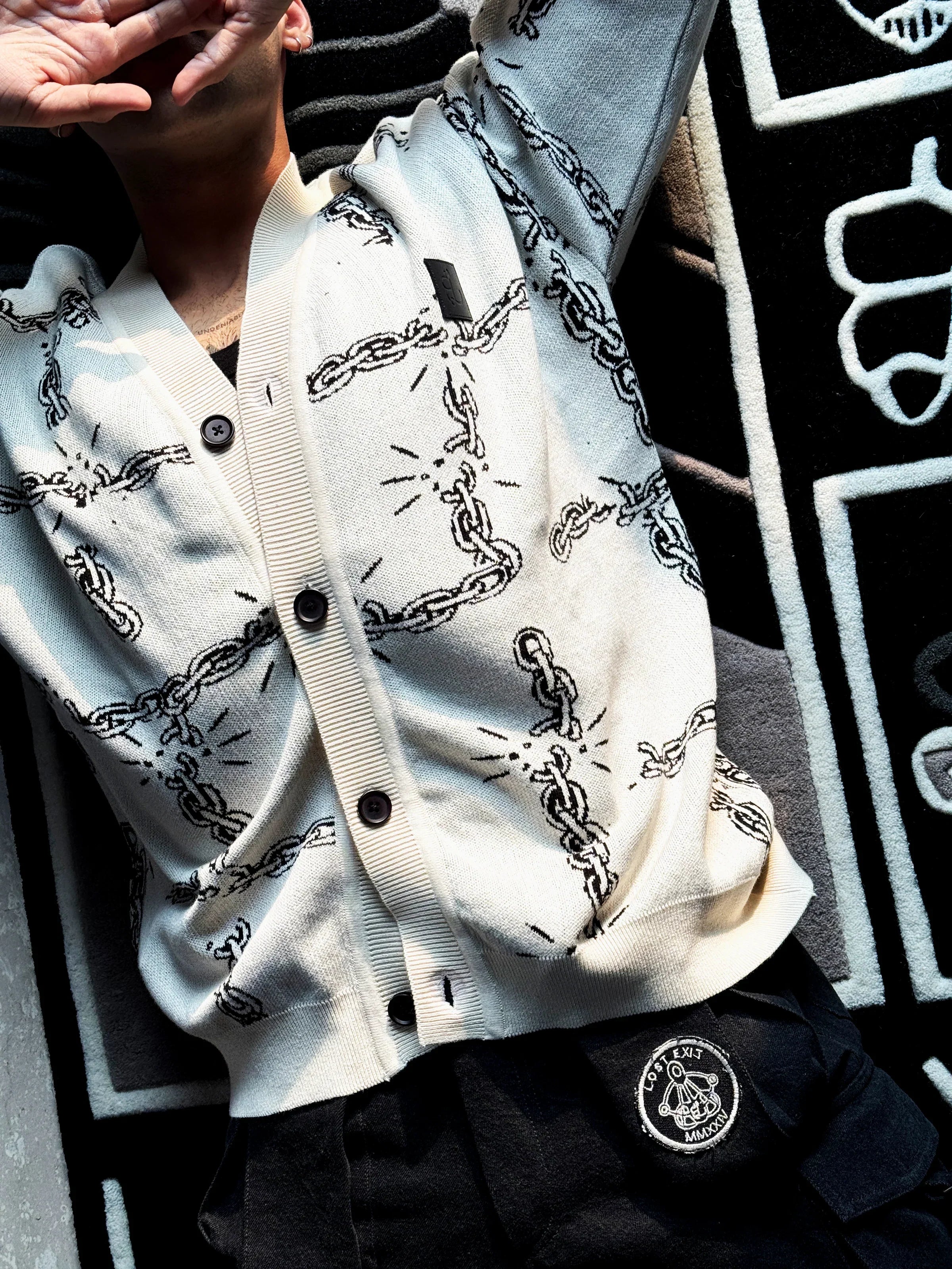 Person wearing a white cardigan with black chain pattern and black pants with a Lost Exit patch lying on a black and white patterned carpet