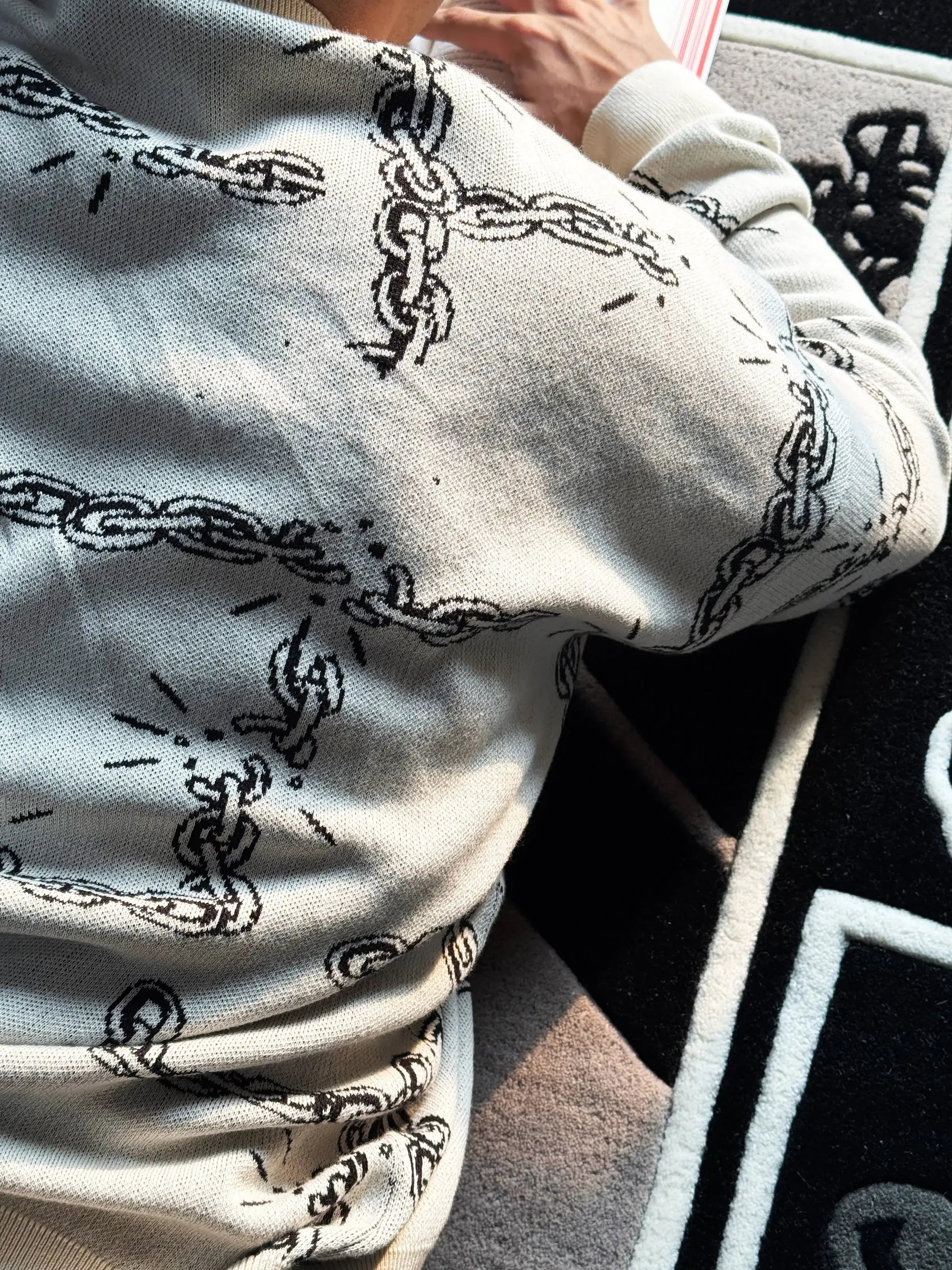 Person wearing light gray sweater with black broken chain graphic design lying on patterned carpet